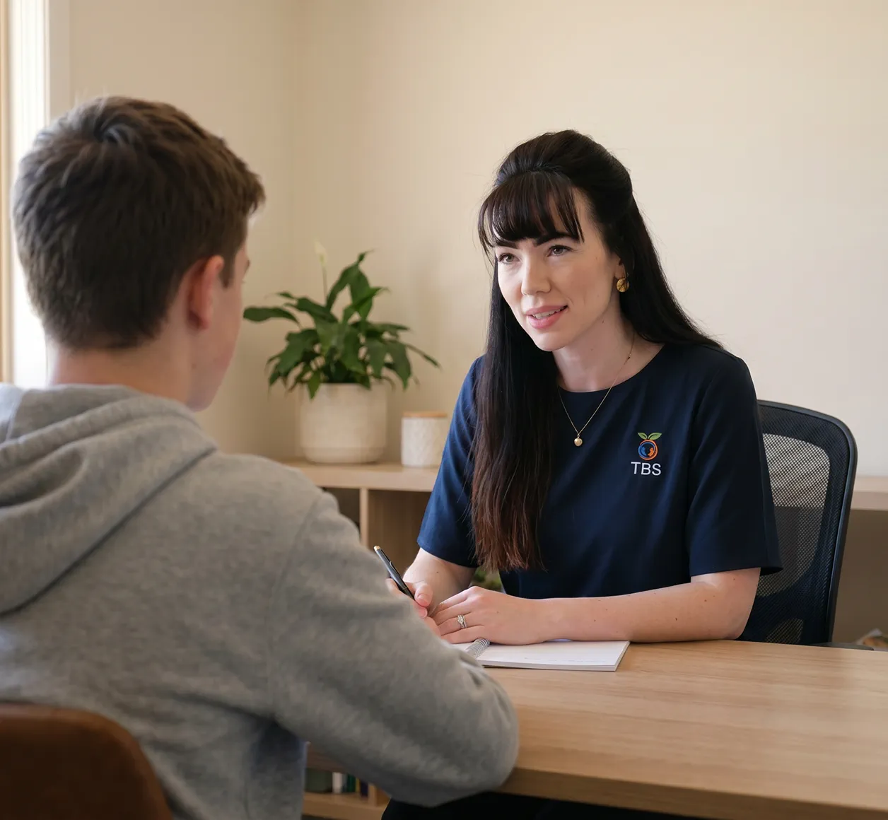 A Target Behaviour Services practitioner sits across a wooden desk speaking with a young participant during a behaviour support session.