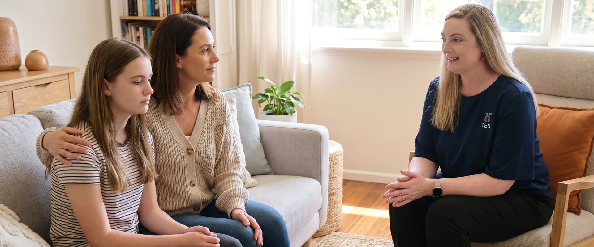 A Target Behaviour Services practitioner speaks with a mother and her teenage daughter on a couch in a sunlit living room.