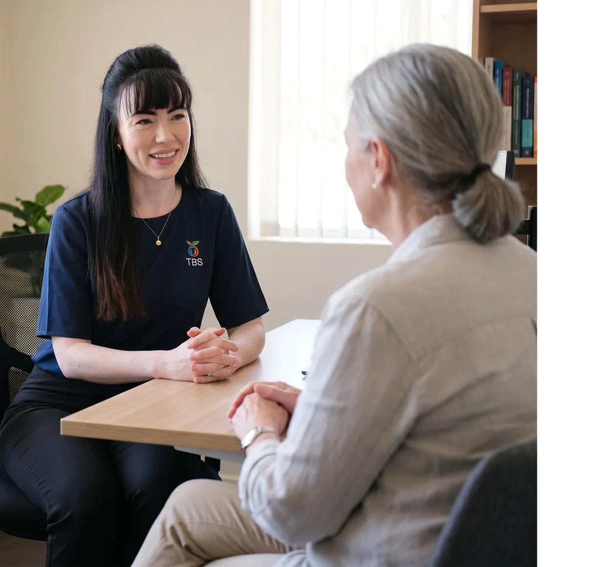 A Target Behaviour Services practitioner smiles warmly while speaking with an older female participant at her desk.
