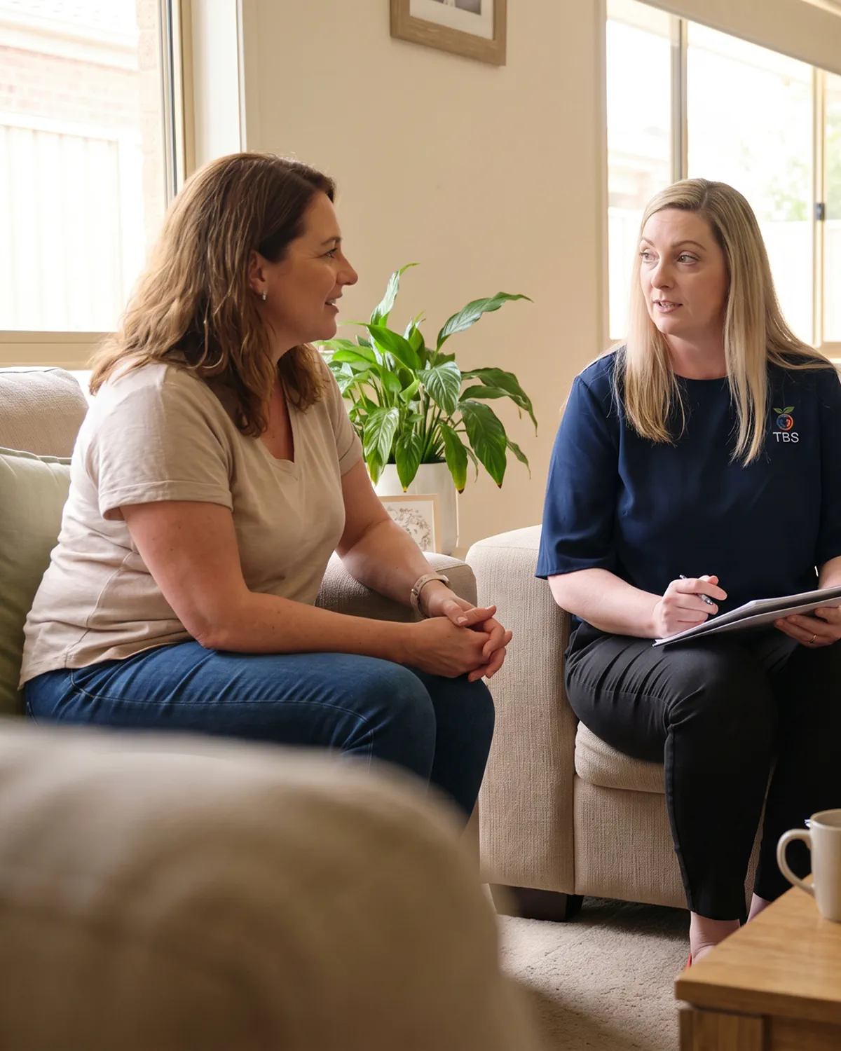 A Target Behaviour Services practitioner sits in a client home, listening attentively while taking notes as a mother shares her concerns.
