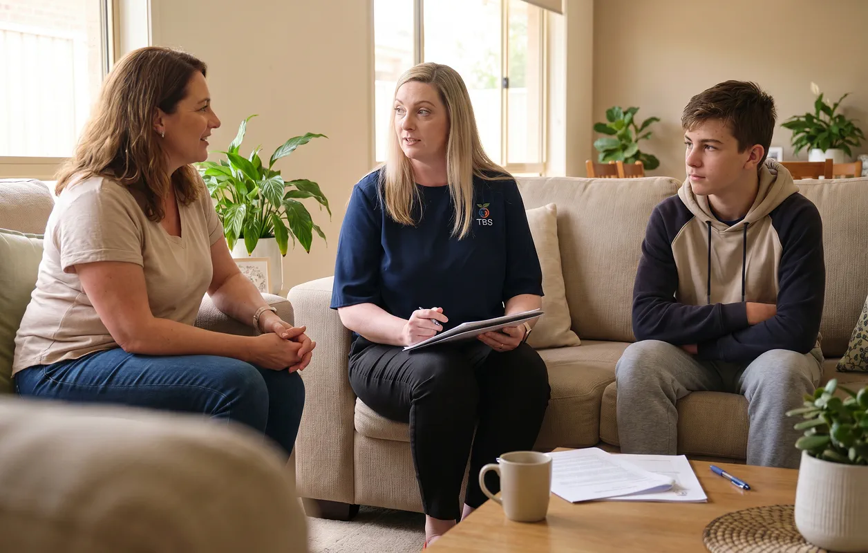 A Target Behaviour Services practitioner sits with a mother and her teenage son in their living room during a behaviour support discussion.