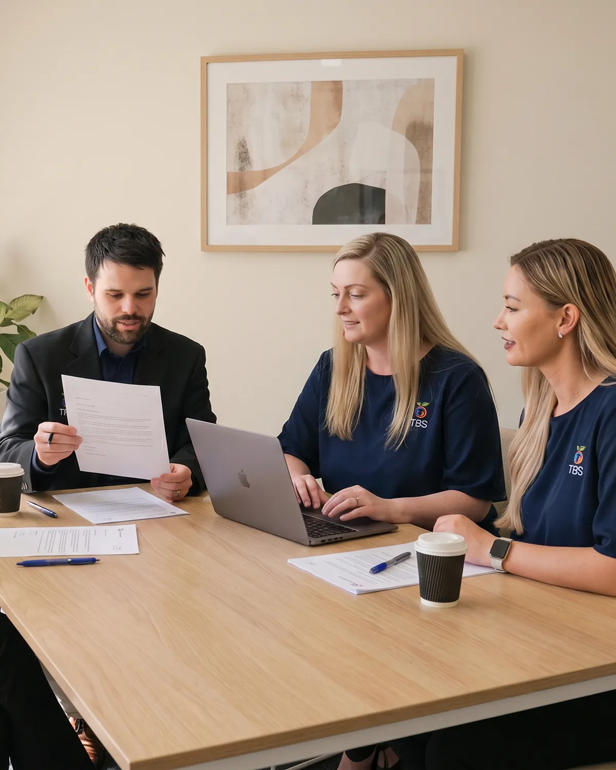 Three Target Behaviour Services team members review documentation together at a meeting table with a laptop and printed notes.