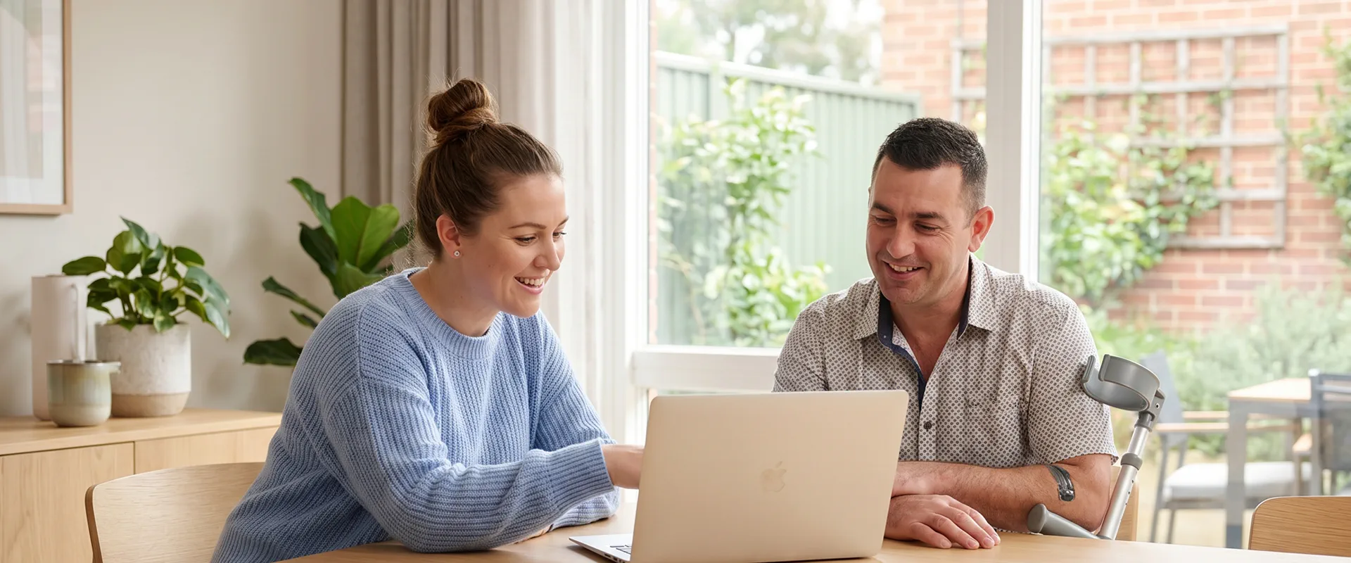 A support coordinator and a participant who uses a forearm crutch review information together on a laptop at a kitchen table.